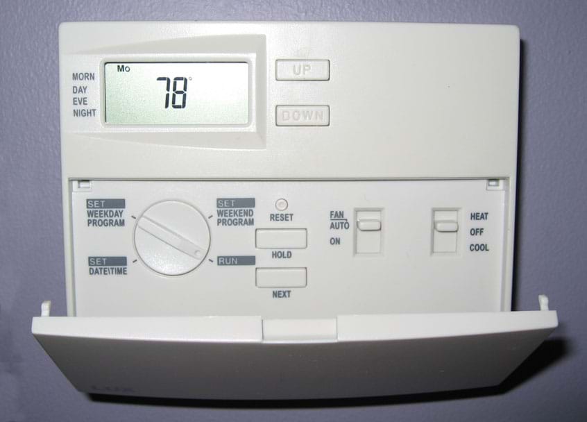 Photo shows an 11 x 9 cm plastic box mounted on a wall, displaying a temperature of 78 °F and many dials and buttons for programming temperature, dates and times.